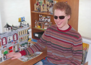 Matthew Shifrin sitting at a table with several completed Lego sets. He has ginger hair and sunglasses on.